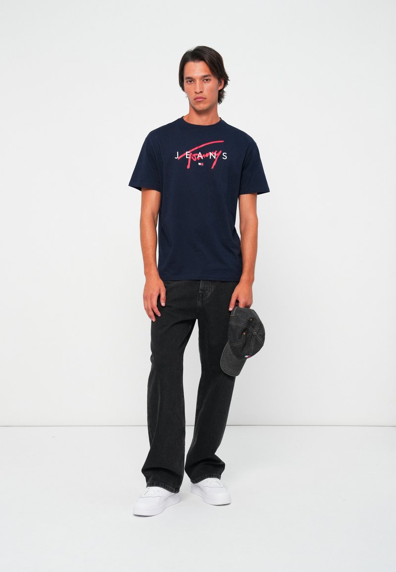 T-shirt en coton bleu marine avec un texte graphique rouge et blanc ; associé à un jean droit noir et une casquette grise, porté avec des baskets blanches.
