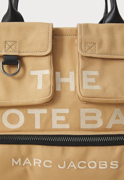 Kangaskassi, jossa on mustat nahkahandjat, kaksi etutaskua, musta vetoketju ja valkoinen teksti "THE TOTE BAG" ja "MARC JACOBS."