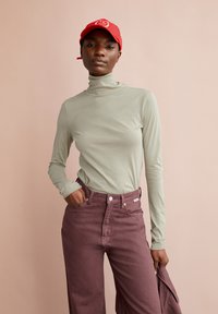 Top a maniche lunghe e collo alto in tessuto verde chiaro. Abbinato a pantaloni larghi a vita alta di colore bordeaux. Il modello tiene in mano una giacca e indossa un berretto rosso.