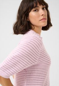 Lyserød strikket sweater med korte ærmer, præget af et tekstureret mønster af åbne sting og en rund halsudskæring.