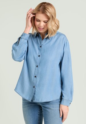 Woman with blonde hair wearing a light blue denim shirt and jeans, touching her hair, looking down, neutral background.