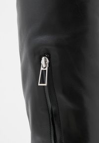 Detailní záběr na stříbrný jezdce zipu na černém koženém povrchu, ukazující podrobnou strukturu a stehy.