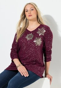 Burgunderfarbener V-Ausschnitt-Pullover mit metallisch-silbernen Schneeflockenmuster, langen Ärmeln und speckled Textur. Getragen mit dunkelblauen Jeans.