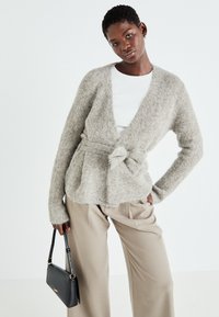 Cardigan tricotat de culoare gri deschis, cu un cordon la talie, decolteu adânc în V, mâneci lungi și o textură moale. Asortat cu pantaloni bej și o geantă neagră.