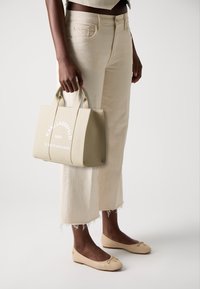 Sac à main beige texturé avec des poignées courtes et un texte imprimé blanc, tenu à côté d'un pantalon large couleur crème avec un ourlet effiloché et des ballerines beiges.