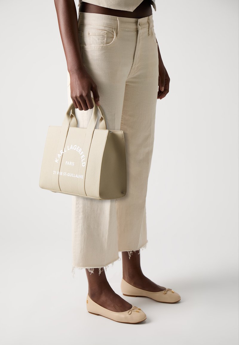 Sac à main beige texturé avec des poignées courtes et un texte imprimé blanc, tenu à côté d'un pantalon large couleur crème avec un ourlet effiloché et des ballerines beiges.