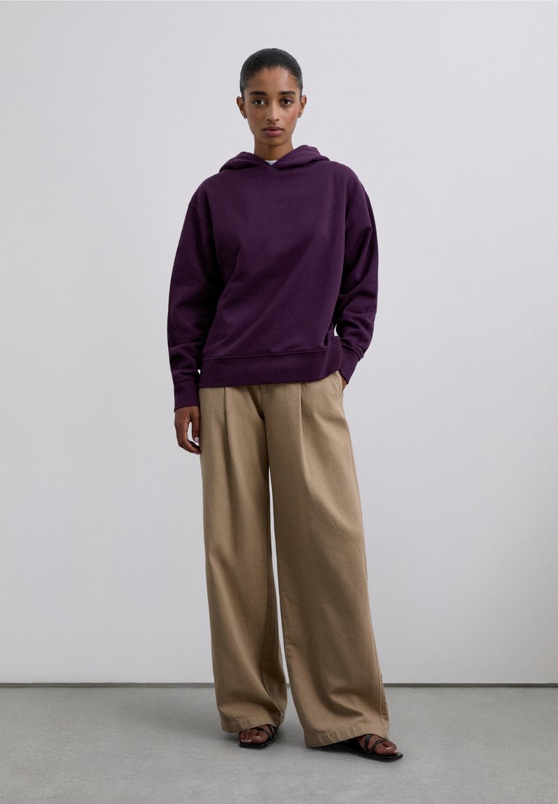 Purpurowa bluza z kapturem ze sznurkiem, o luźnym kroju i ściągaczach na rękawach, zestawiona z szerokimi beżowymi spodniami i czarnymi sandałami.