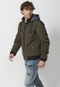 Giacca zip-up verde oliva con cappuccio grigio, presenta più tasche frontali, polsini elasticizzati e una finitura testurizzata. Stile casual.