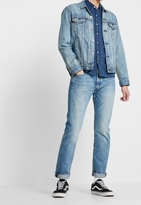 Muž oblečený v světle modré džínové bundě a džínách, tmavě modré košili a černo-bílých teniskách, stojící s jednou rukou v kapse.