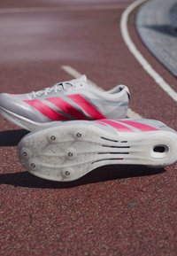 Harmaat urheilukengät kirkkaan vaaleanpunaisilla korostuksilla ja kolmella raidalla. Omistaa teksturoidun pohjan piikkien kanssa ja virtaviivaisen muotoilun tehokkuutta varten.