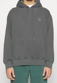 Uomo che indossa una felpa Carhartt grigio scuro sbiadita con tasca frontale e pantaloni verdi, mostrato dalla collottola ai fianchi.
