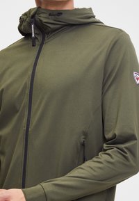 Giacca leggera verde oliva con cappuccio, dalla texture liscia, con zip frontale completa e tirazip nera; presenta un logo sulla manica sinistra.
