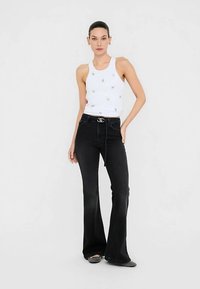 Witte tanktop met kleine gouden versieringen, gecombineerd met hoog taille zwarte uitlopende jeans en zwarte schoenen; eenvoudig ontwerp en pasvorm.