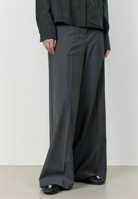 Gray wide-leg trousers made of smooth fabric, featuring a high waist and sleek cut. Paired with a dark, slightly sheer, long-sleeve shirt.