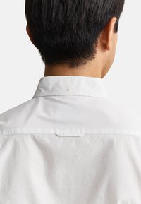 Cabello corto y negro visible desde atrás en una persona que lleva una camisa blanca con cuello y un botón en el cuello.
