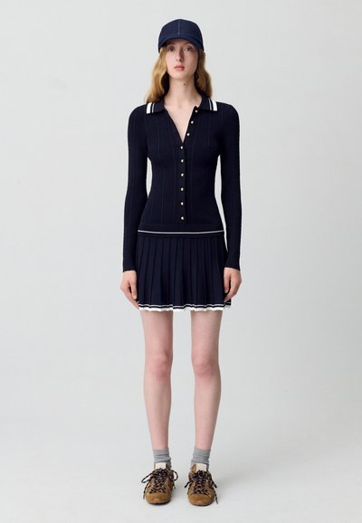 Abito in maglia blu navy con maniche lunghe, bottoni dorati e colletto a righe bianche; sezione gonna plissè; abbinato a sneakers marroni e calze grigie.