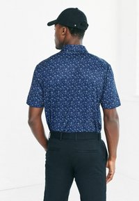 T-paidassa on lyhyet hihat, tummansininen väri ja teksturoitu, hajautettu valkoinen kuvio; yhdistetty mustiin housuihin. Säädettävä musta lippalakki näkyy.