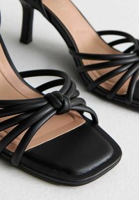 Czarne sandały na obcasie mają kwadratowy kształt noska, węzłowe skórzane akcenty i wiele nachodzących na siebie pasków. Wnętrze ma beżowe wykończenie.