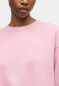 Κοντινό πλάνο ενός ατόμου που φοράει ανοιχτό ροζ φούτερ με μικρό λευκό λογότυπο Jumpman στο στήθος.