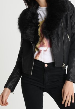Femme portant une veste en cuir noire avec un grand col en fourrure noire, un t-shirt blanc à motif graphique et un pantalon noir taille haute.