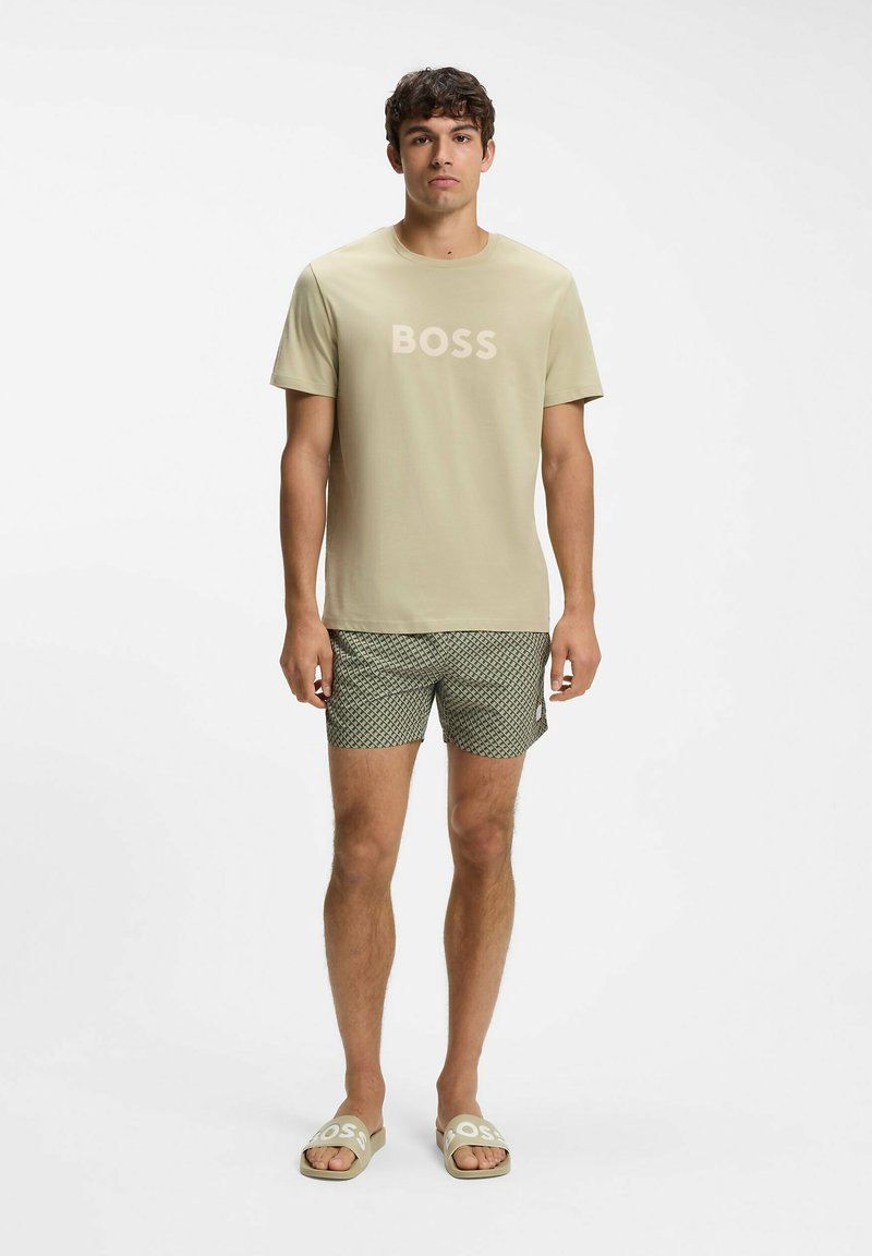 Jeune homme debout portant un t-shirt beige "BOSS", un short à motifs et des sandales beiges assorties avec le logo "BOSS" sur fond blanc uni.