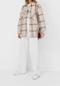 Rutig flanellskjorta i beige, krämfärgade och bruna nyanser, med två bröstfickor, knäppningar och långa ärmar över vita vida byxor.