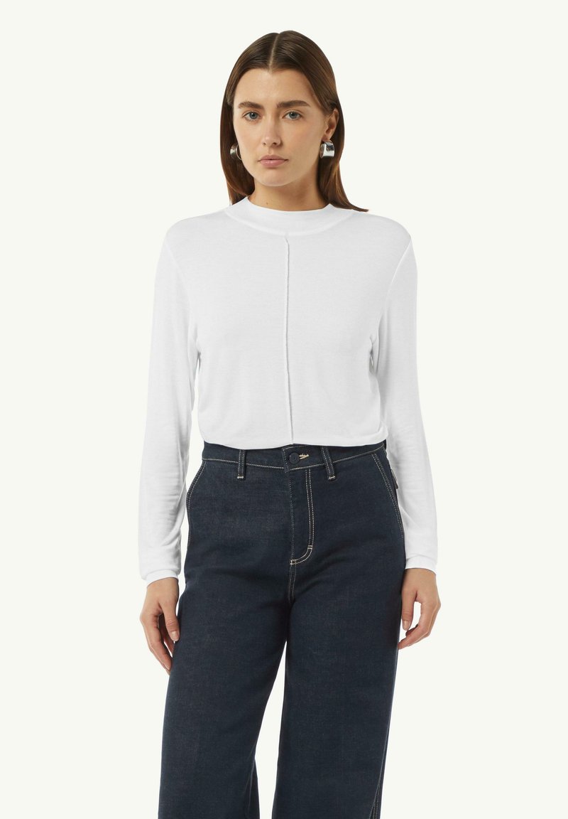 Weißes Langarmoberteil mit hohem Kragen und zentraler Nahtdetail, kombiniert mit hochgeschnittenen, dunklen Jeans. Glatte Textur, figurbetontes Design.