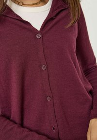 Cardigan bordeaux avec un design à l'avant à boutons, manches longues, texture douce et capuche. Boutons violets remarquables à l'avant.