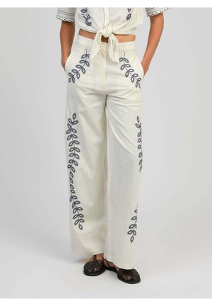 Femme portant un pantalon blanc taille haute à jambes larges avec une broderie de feuilles noires et des poches, associé à des sandales noires tissées.