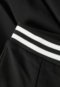 Melnas auduma treneris bikses ar teksturētu jostasvietu, kurā ir divas horizontālas baltas svītras. Gluda virsma un brīvs pieguļojums ar sānu kabatām.