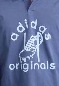 Ljubičasta pamučna majica s v-izrezom, s bijelim grafičkim prikazom fudbalske kopačke i napisom "adidas originals" u zaobljenim slovima.