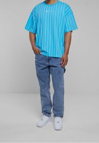 Henkilö, joka on pukeutunut siniraidalliseen lyhythihaiseen paitaan, löysi sinisiin farkkuihin ja valkoisiin tennareihin, seisoo tasaisen harmaan taustan edessä.