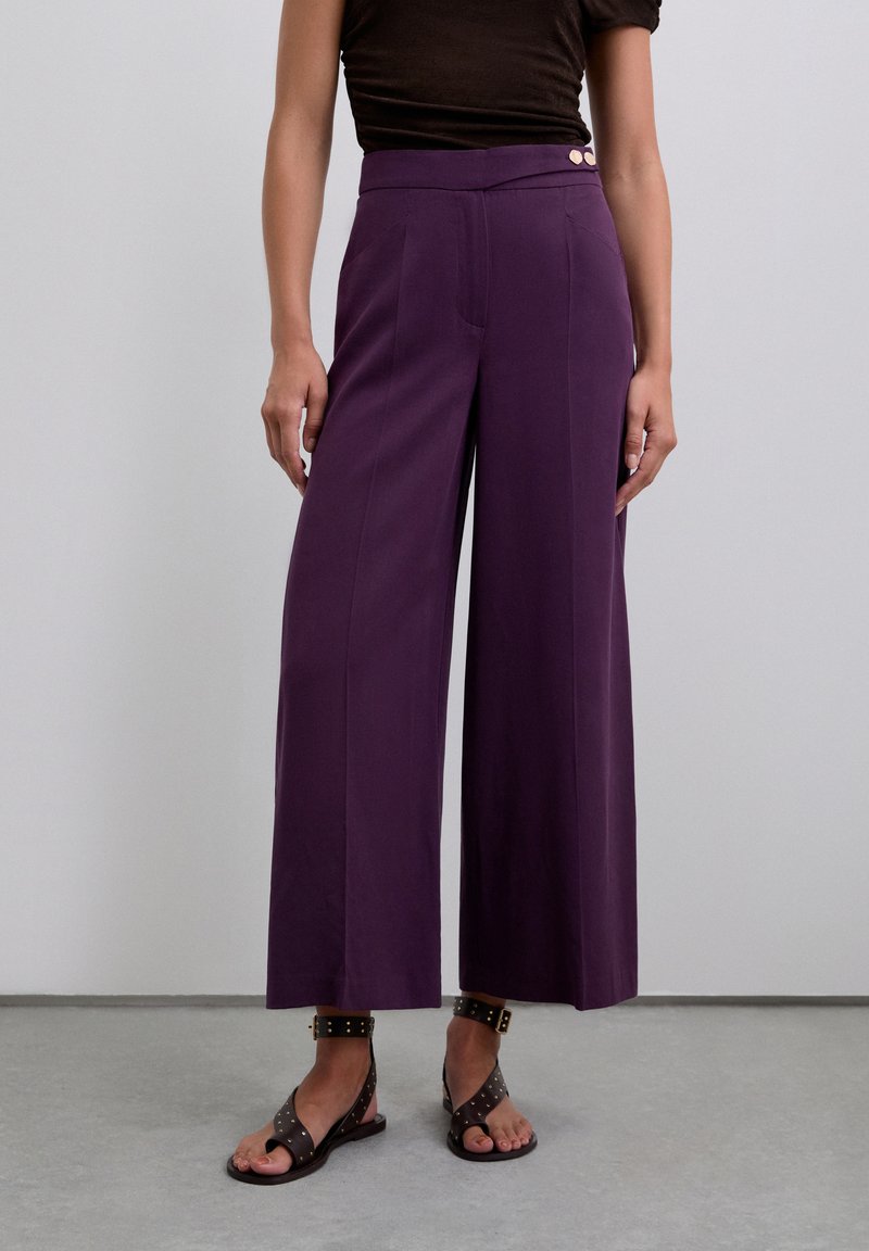 Pantalons larges violets avec une texture lisse, dotés d'une taille haute et de deux boutons sur le côté. Portés avec des sandales noires à clous.