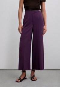 Wide-leg purple trousers with a smooth texture, featuring a high waistband and two buttons on the side. Worn with dark, studded sandals.