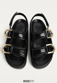 Svarte patent sandaler med justerbare stropper, guldspænder, flade såler og en rund form. Glat tekstur med åbent tådesign.