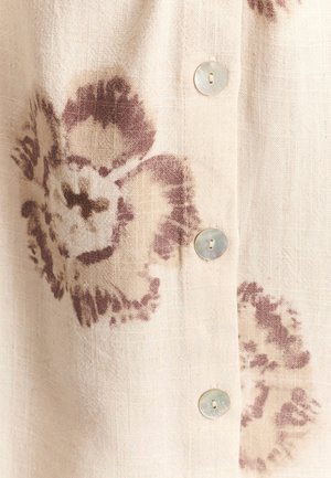 Blouse en lin crème imprimée de fleurs marron foncé. Elle comporte des boutons transparents sur le devant et un tissu à texture.