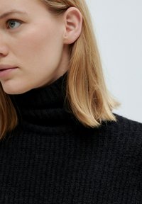 Zwarte gebreide coltrui met een textuurpatroon en geribde kraag, gedragen door een model met schouderlang blond haar.