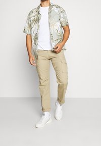 Mies kävelee valkoisessa t-paidassa, vihreässä kukallisessa lyhythihaisessa paidassa, beigeissä cargo-housuissa, valkoisissa sukissa ja valkoisissa Adidas-tenniskenkässä.