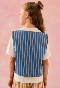 Denim prsluk s vertikalnim bijelim prugama, s okruglim izrezom i kontrastnim šavovima, nošen preko bijele košulje s kratkim rukavima.