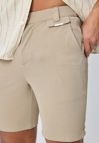 Shorts beiges en tissu texturé avec un devant plat, des poches latérales, des passants de ceinture et un accent métallique argenté au niveau de la taille.