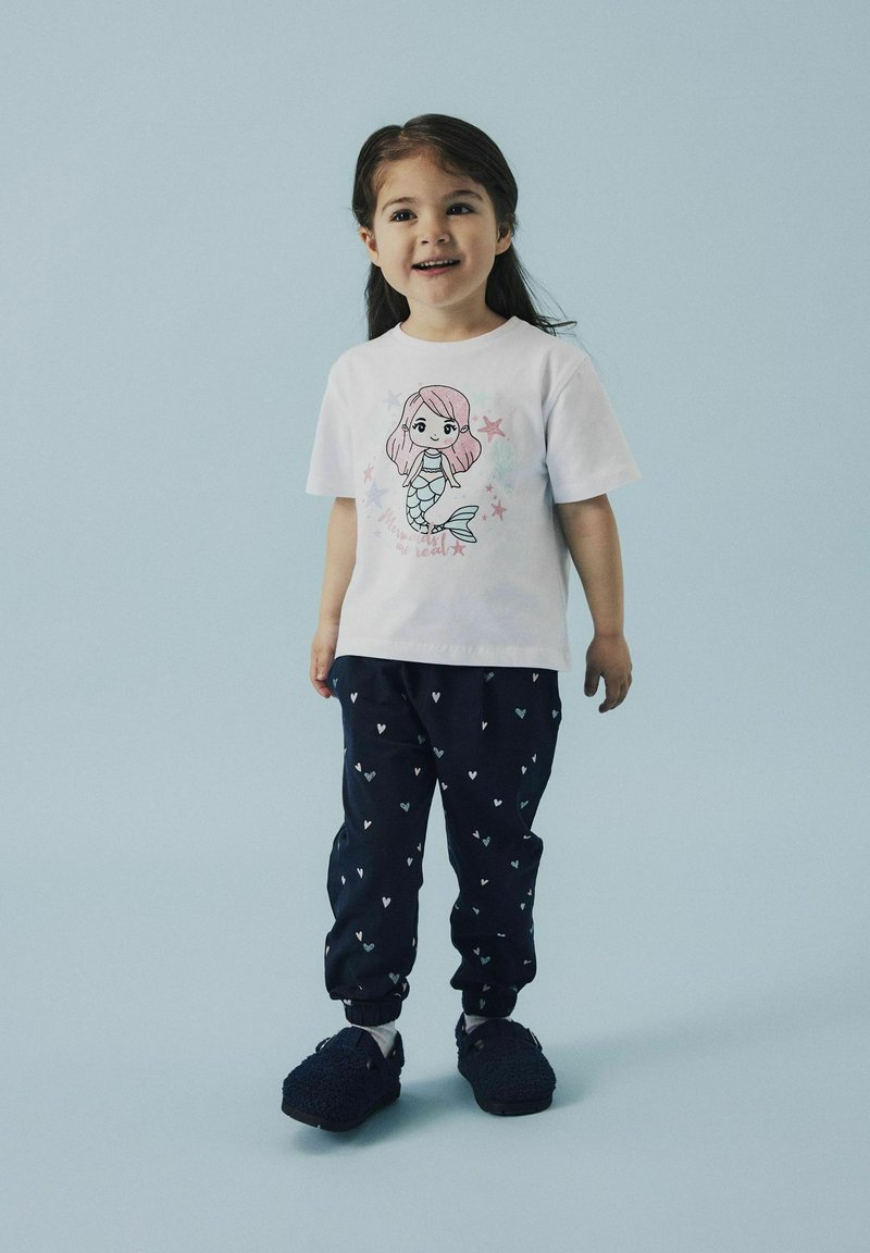 Niña con una camiseta blanca con un gráfico de sirena, pantalones navy con pequeños corazones y pantuflas oscuras y peludas contra un fondo claro.