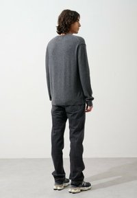 Persona con capelli scuri di lunghezza media che indossa un maglione grigio, jeans neri e sneakers nere, in piedi e che guarda di lato su uno sfondo semplice.