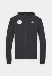 Crna zip-up dukserica s kapuljačom i vezicom, s kružnom zelenom naljepnicom s dizajnom nogometne lopte i bijelim Adidas logom na prsima.