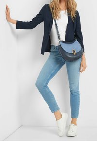 Blaue und marineblaue Leder-Umhängetasche mit geschwungener Form, goldener Hardware und einer strukturierten Wildlederklappe, getragen zu hellblauen Jeans und weißen Sneakers.