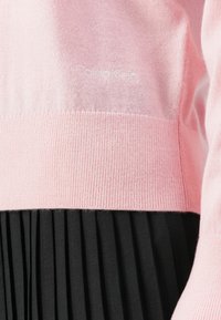 Różowy sweter z dzianiny marki Calvin Klein w połączeniu z czarną spódnicą plisowaną, ukazujący obszar pasa i część rękawa.