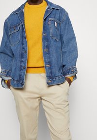Jeansjacke in Blau mit zwei Brusttaschen und Knopfverschluss, getragen über einem gelben Strickpullover. Beige Hosen runden das Outfit ab.