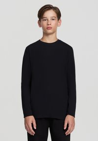 Czarny longsleeve z długim rękawem, wykonany z miękkiego materiału, z okrągłym dekoltem i prostym wykończeniem. Modelka stoi z rękami wzdłuż ciała na neutralnym tle.