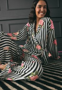 Pajama set s uzorkom prugica u crnoj i bijeloj boji s ružičastim cvjetnim naglasima. Mekani materijal, duži rukavi i detalj s vezanjem u struku. Pozadina s crno-bijelim prugama.