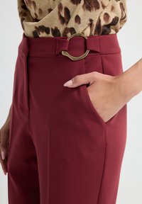 Woman wearing burgundy pants with a fabric belt and gold buckle, leopard print top tucked in, hand in pocket, against a white background.