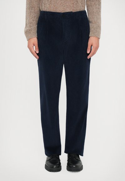 Person, der eine marineblaue Cordhose, schwarze Lederschuhe und einen beigen Strickpullover trägt, steht vor einem schlichten weißen Hintergrund.